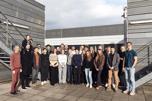 Auf einer Terrasse der Hochschule Düsseldorf steht eine Gruppe von etwa 20 Studierenden für ein Gruppenfoto. Im Hintergrund ist die markante silberne Metallfassade zu sehen.